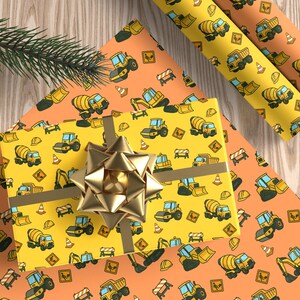 May include: A wrapped gift with a gold bow sits on orange and yellow wrapping paper. The paper is patterned with cartoon construction vehicles, including cement mixers, excavators, and dump trucks. A sprig of evergreen is in the corner.