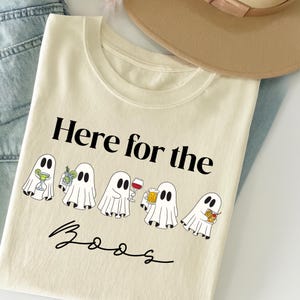 Könnte beinhalten: Cremefarbenes T-Shirt mit dem Text "Here for the Boos" und Cartoon-Geistern, die Cocktails, Wein und Bier halten. Das Shirt ist gefaltet, mit einem beigen Hut und blauen Jeans im Hintergrund.