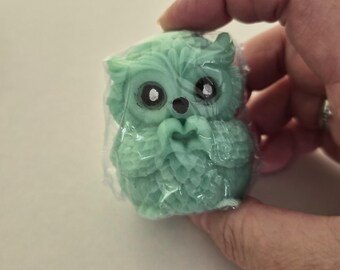 May include: A small, light green owl-shaped object with detailed feather textures, large dark eyes, and a small beak, encased in a clear plastic wrapper. The owl is depicted with its wings folded in front of its chest, creating a heart-like shape.