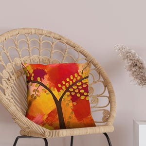 May include: A decorative throw pillow with an abstract tree design in shades of red, orange, and yellow. The pillow is placed on a woven wicker chair. A white vase with dried flowers sits on a white cube.