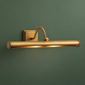 Könnte beinhalten: Eine Messing-Bildleuchte mit rechteckiger Wandhalterung und gebogenem Arm. Die Leuchte hat einen zylindrischen Schirm und strahlt ein warmes Licht aus. Die Leuchte ist an einer dunkelgrünen Wand montiert.