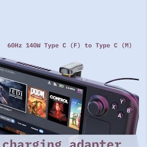 May include: A charging adapter for the Steam Deck. The adapter is silver and connects to the device. The screen displays game titles. Text on the screen reads "Jedi Fallen Order", "Doom Eternal", "Control", and "No Man's Sky". The text "charging adapter for STEAM DECK" is also visible.