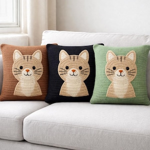 Puede incluir: Tres cojines cuadrados de ganchillo con caras de gato. Los cojines son de color marrón, negro y verde. Cada cojín presenta una cara de gato color canela con bigotes negros, una nariz rosa y rayas marrones. Los cojines están sobre un sofá blanco.