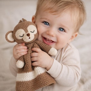 Könnte beinhalten: Ein gehäkeltes Affenspielzeug in Braun und Creme mit geschlossenen Augen und einem lächelnden Gesicht. Das Spielzeug hat einen runden Kopf, Ohren und Gliedmaßen. Der Affe wird von einem lächelnden Baby mit blauen Augen gehalten.