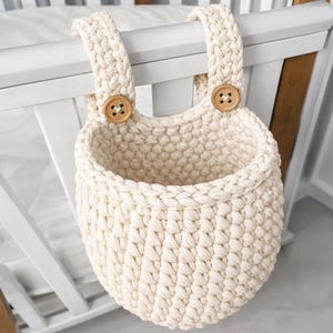 Pode incluir: Cesto de berçário de crochê creme, com duas alças tecidas e fechos de botão de madeira. O cesto foi projetado para pendurar em um berço ou estrutura semelhante, proporcionando armazenamento conveniente.