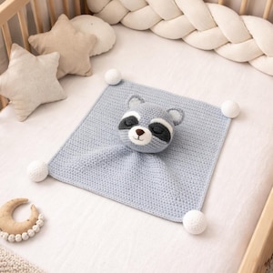 Puede incluir: Una manta de seguridad para bebé de ganchillo azul claro con una cara de mapache y pompones blancos. La manta está en una cuna de madera con almohadas en forma de estrella y un mordedor de luna de madera.