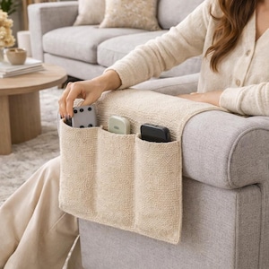 May include: A cream-coloured armrest tidy with three pockets, holding a remote control, a small device, and a black remote. The organiser is on the arm of a grey sofa. The scene is in a living room.