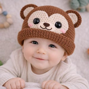 Może przedstawiać: Brązowo-beżowa szydełkowa czapeczka dla dziecka z motywem małpiej twarzy. Czapeczka ma duże czarne oczy, różowe policzki i małe uszy. Dziecko ma na sobie białą koszulkę z długim rękawem.