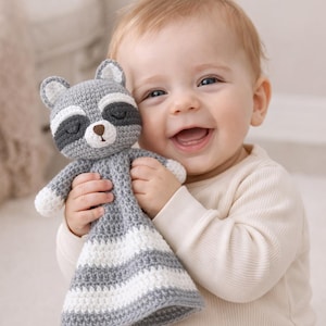 Może przedstawiać: Szara i biała szydełkowa kołderka z szopem praczem. Szop ma szarą maskę, uszy i biały pasek. Kocyk ma naprzemienne szare i białe paski.