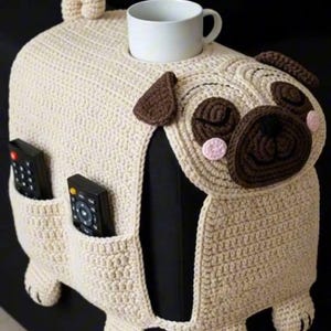 Puede incluir: Una funda para reposabrazos de sofá con forma de pug, de color crema y crochet, con un portavasos y dos bolsillos. El pug tiene orejas, ojos y nariz marrones, con detalles en rosa. Dos mandos a distancia están en los bolsillos. Una taza blanca está en el portavasos.