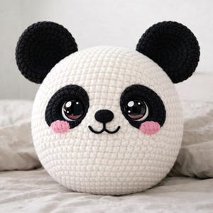 May include: A round, white crocheted panda pillow with black ears, eyes, and eye patches. The panda has pink cheeks and a small black nose and mouth. The pillow is sitting on a white surface.