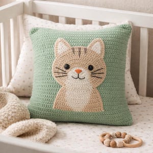 May include: A square, crocheted cushion featuring a cat face design. The cushion has a light green background with a tan and cream-coloured cat face in the centre. The cat has black whiskers, a small orange nose, and a smiling mouth. The cushion is on a white cot.