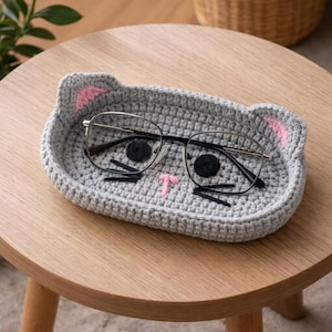 May include: A gray crocheted cat-shaped tray with pink ear details and a stitched face, holding a pair of eyeglasses. The tray is on a wooden table, showcasing a handmade item for home organization and decor.