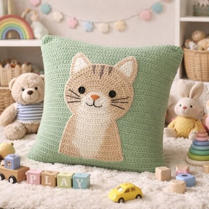 May include: A green crochet cushion featuring a tan cat design. The cat has black eyes, whiskers, and a pink nose. The square cushion is on a white rug, surrounded by toys such as a teddy bear, a toy car, and blocks spelling "PLAY".