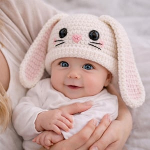 Pode incluir: Um bebê usando um chapéu de coelho de crochê branco com orelhas internas rosa, olhos pretos e bigodes. O bebê tem olhos azuis e está usando uma camisa branca de manga comprida. O chapéu é um acessório fofo.