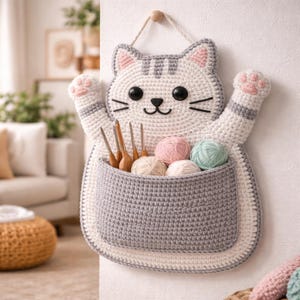 May include: A handmade, crochet cat-shaped wall organizer in white, gray, and pink. The cat has black eyes, whiskers, and a gray striped head. The pocket holds crochet hooks and yarn balls in various colors. It hangs from a wooden peg.