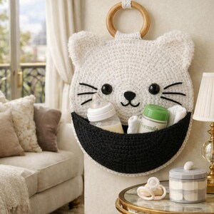 May include: A white crochet cat-shaped wall organizer with a black pocket, holding a baby bottle, a container, and a rolled-up cloth. The cat has black eyes and whiskers, and a wooden ring for hanging.
