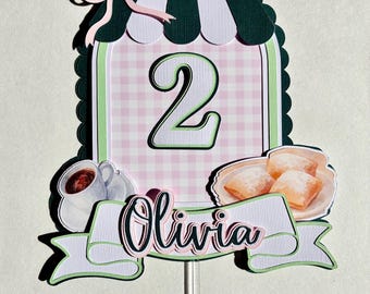 Pode incluir: Topper de bolo de aniversário de dois anos com um padrão xadrez rosa e branco, um design de toldo verde e branco e o nome "Olivia". O design inclui uma xícara de café, beignets e um laço rosa.