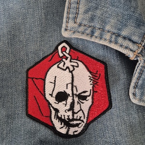 May include: Embroidered patch with a white skull design on a red hexagonal background, attached to a light blue denim jacket. The patch has a half-face design, and the jacket's details are visible.