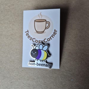 Peut inclure: Épinglette argentée en forme d'abeille avec des ailes violettes, jaunes et bleues, et le texte "non-beenary". L'épinglette est sur une carte blanche avec un logo de tasse à café et le texte "TeesCosyCorner".