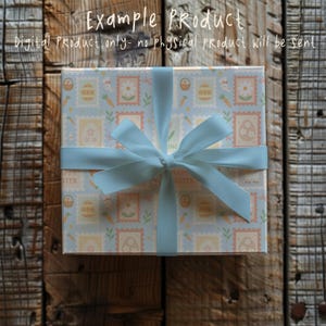 May include: A wrapped gift box with a light blue ribbon and bow. The box is covered in a light blue paper with Easter-themed stamps, including eggs, carrots, and flowers. The text "Example Product" and "Digital Product only- no physical product will be sent" are visible.