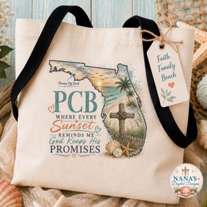 Könnte beinhalten: Eine cremefarbene Canvas-Tragetasche mit schwarzen Trägern, bedruckt mit der Umrisskarte von Florida, einem Strand-Sonnenuntergang, einem Holzkreuz und Muscheln. Der Text lautet: PCB WHERE EVERY SUNSET REMINDS ME GOD KEEPS HIS PROMISES. Mit Anhänger: Faith Family Beach.