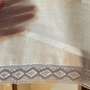 Könnte beinhalten: Ein halbtransparenter weißer Vorhang mit einer dezenten gewebten Gitterstruktur und einer dekorativen weißen Spitzenborte am unteren Rand. Der Stoff ist lichtdurchlässig und die Spitze zeigt ein sich wiederholendes geometrisches Muster.
