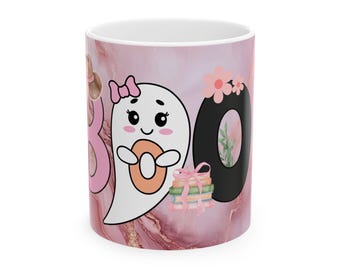 Puede incluir: Taza de cerámica blanca con diseño de mármol rosa y detalles dorados. La taza presenta un fantasma de dibujos animados sosteniendo una rosquilla, la palabra "BOO" en rosa y el número "0" en negro con flores rosas. Una pila de libros está atada con un lazo rosa.