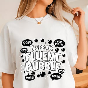 May include: White t-shirt with the text "I Speak Fluent Bubble" in a playful, bubble-like font. The design includes speech bubbles with words like "Pop!", "Yay!", "More!", and "Again!" in black. The shirt is a casual, everyday wear item.