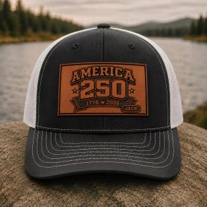 May include: A charcoal grey and white trucker hat featuring a rectangular brown leather patch on the front. The patch is engraved with the text AMERICA 250, the years 1776 and 2026, stars, and the name JACK in a small separate box at the bottom.