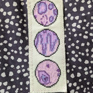 May include: A rectangular, off-white, cross-stitched bookmark featuring three circular designs. Each circle is outlined in black and filled with shades of purple and pink. The bookmark is placed on a dark background with white polka dots.