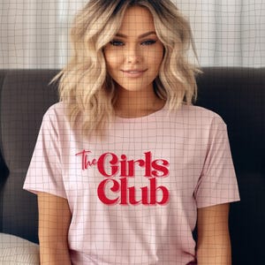 Può includere: Una maglietta rosa chiaro con la scritta "The Girls Club" in rosso. La maglietta ha maniche corte e scollo tondo. Il testo è in un carattere audace e stilizzato.
