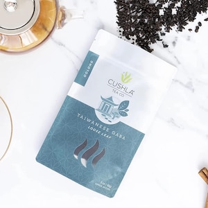 Könnte beinhalten: Eine Packung mit losem Blatt-Tee, Taiwanese Gaba Oolong, von Cushla Tea Co. wird auf einer weißen Oberfläche gezeigt. Die Packung ist weiß und blaugrün mit dem Text "Oolong" und "Taiwanese Gaba Loose Leaf". Eine Glaskanne und eine Tasse sind ebenfalls zu sehen.