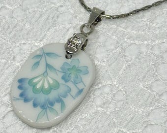 Pode incluir: Colar prateado com um pendente oval. O pendente apresenta um design floral azul e verde sobre um fundo branco. A corrente é fina e o fecho do pendente tem um pequeno detalhe brilhante. O design floral lembra porcelana vintage.