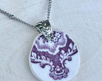 May include: A silver-toned necklace with a pendant featuring a white background and a purple floral design. The pendant is oval-shaped and has a decorative silver clasp. The chain is a delicate beaded style.