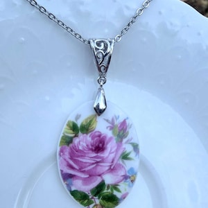 May include: An oval-shaped pendant featuring a detailed illustration of a large pink rose with green leaves and small blue flowers. The pendant is attached to a silver-toned metal bail and a matching silver-toned chain necklace.