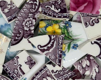 May include: A collection of broken ceramic pieces with floral and patterned designs. The fragments feature a mix of colors, including white, purple, yellow, and blue. Some pieces display intricate floral patterns, while others have decorative borders.