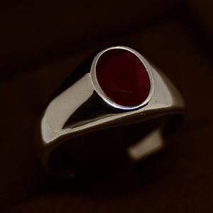 May include: A silver ring with a polished finish, featuring a deep red, oval-shaped stone. The ring is set in a classic bezel setting, presented in a brown jewelry box. The ring's design is simple and elegant.