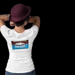 May include: White t-shirt featuring a white convertible car graphic on a candy bar wrapper with the text "FUN-SIZED". The person is wearing a burgundy hat and blue jeans. The background is black.
