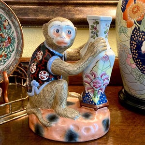 May include: A ceramic monkey figurine holding a decorative vase. The monkey is wearing a black vest with red and white circular designs. The vase is white with floral patterns and a blue base. The monkey is sitting on a peach-colored base with black spots.