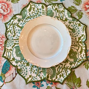 May include: A white ceramic plate with a gold rim sits atop a green and white floral placemat. The placemat has a scalloped edge and is placed on a floral tablecloth with pink and green flowers.