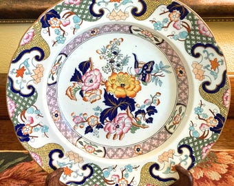 May include: A decorative ceramic plate with a floral design. The plate features a white background with a central floral arrangement in pink, yellow, and blue. The rim has a scalloped edge with a repeating pattern of flowers and geometric shapes.