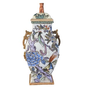 May include: A decorative ceramic vase with a lid, featuring a detailed hand-painted design. The vase showcases colorful birds, floral patterns, and a square base. The handles are a gold color, and the lid is topped with a small, brown figurine.