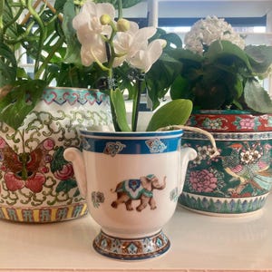 May include: A white ceramic planter with a blue rim and an elephant design. The planter has two small handles and is filled with a white orchid. Two other decorative planters are in the background.