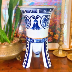 May include: A white and blue ceramic vase with a Greek key design and floral accents. The vase has a flared top with a gold rim and is supported by three legs. The vase is approximately 12 inches tall.