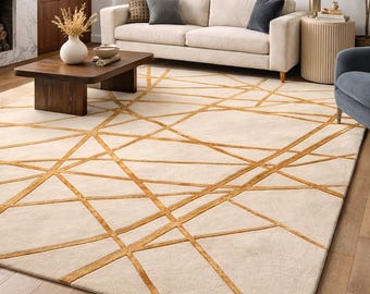 May include: A cream-colored area rug with a geometric pattern of intersecting gold lines. The rug is placed on a hardwood floor in a living room setting, with a sofa, coffee table, and artwork visible in the background.