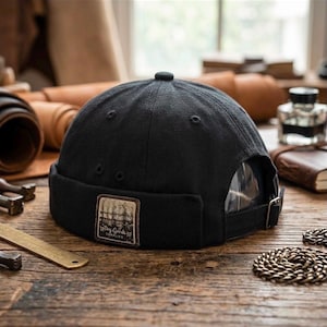 Puede incluir: Una gorra negra sin visera con borde enrollado y un parche rectangular marrón en la parte posterior. El parche muestra una ilustración gráfica de una valla y el texto Dry Goods SUPPLIES. La gorra incluye una correa con hebilla metálica.