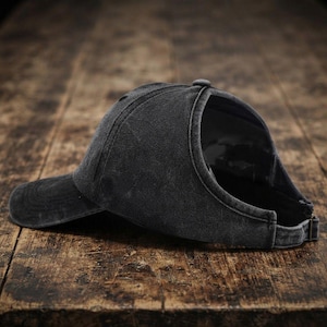 May include: A charcoal grey baseball cap with a distressed, washed-out finish. The cap features a curved brim and a large, open cutout at the back designed for a ponytail, secured with an adjustable metal buckle strap at the base of the crown.