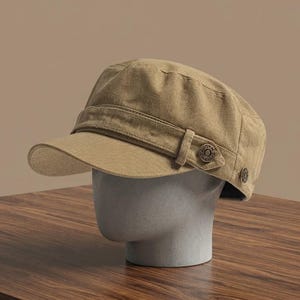 Puede incluir: Una gorra de estilo militar color canela con una corona estructurada, una visera corta y rígida, y una banda horizontal en la parte delantera. La banda está asegurada por dos botones metálicos circulares con detalles en relieve en los lados.