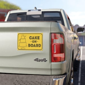 Puede incluir: Un letrero rectangular amarillo con texto negro y una ilustración de pastel está adherido a la parte trasera de una camioneta verde claro. El letrero dice "CAKE -ON- BOARD" y presenta un diseño de pastel de tres niveles.